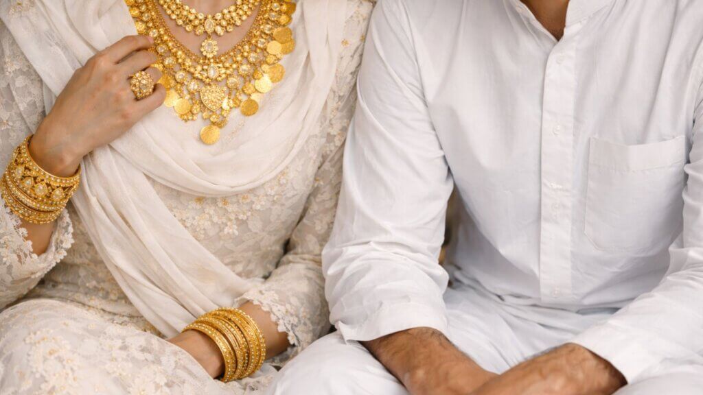 Muslim woman wearing traditional white attire adorned with gold jewelry sitting beside a man dressed in simple white clothing, illustrating the Islamic rule permitting gold for women but not for men.